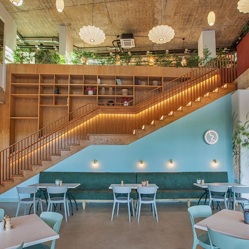 Cozy modern cafe interior with wooden staircase and turquoise seating area, stylish lighting and shelving for a relaxing dining experience.